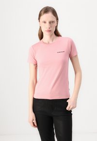 Rózsaszín, testhezálló pamut póló, rövid ujjakkal és kerek nyakkivágással. Fekete "GUESS JEANS" logó nyomtatva a mellkason.