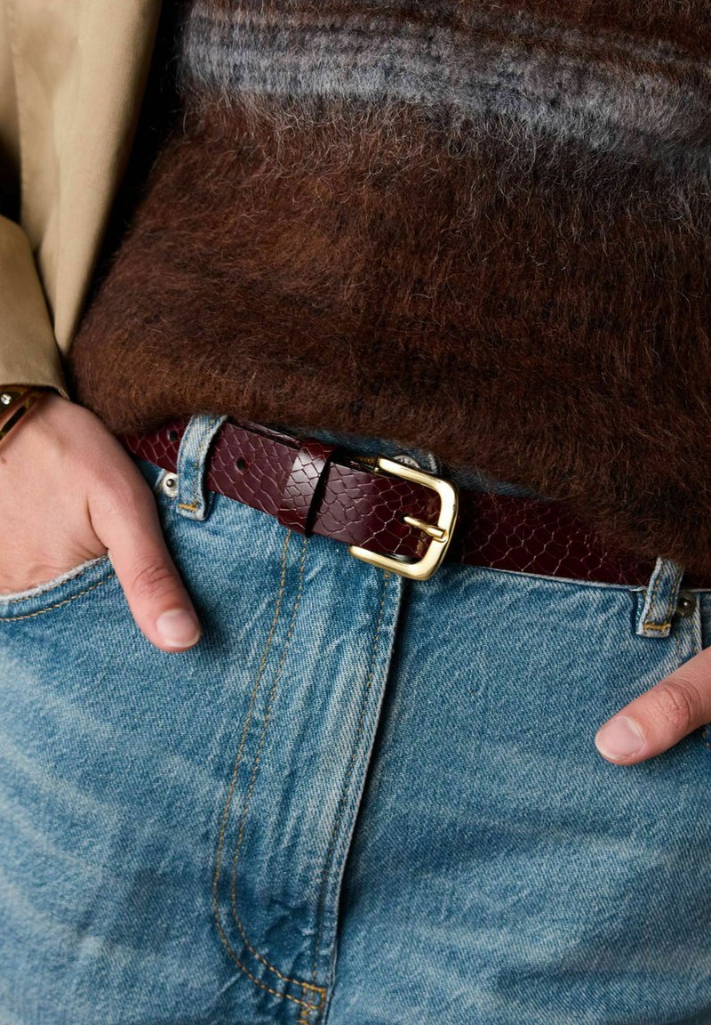 Persoon gekleed in blauwe jeans en een bruine donzige trui met handen in de zakken, accessorized met een donkerrode gestructureerde leren riem met gouden gesp.