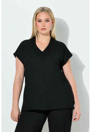 Blonde Frau, die ein schwarzes V-Ausschnitt-Oberteil mit kurzen Ärmeln und passende schwarze Hosen trägt, steht vor einer weißen Wand.