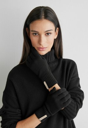Gants en tricot noir avec poignets côtelés, dotés d'une étiquette au niveau du poignet. Portés par une personne en pull foncé sur un fond neutre.
