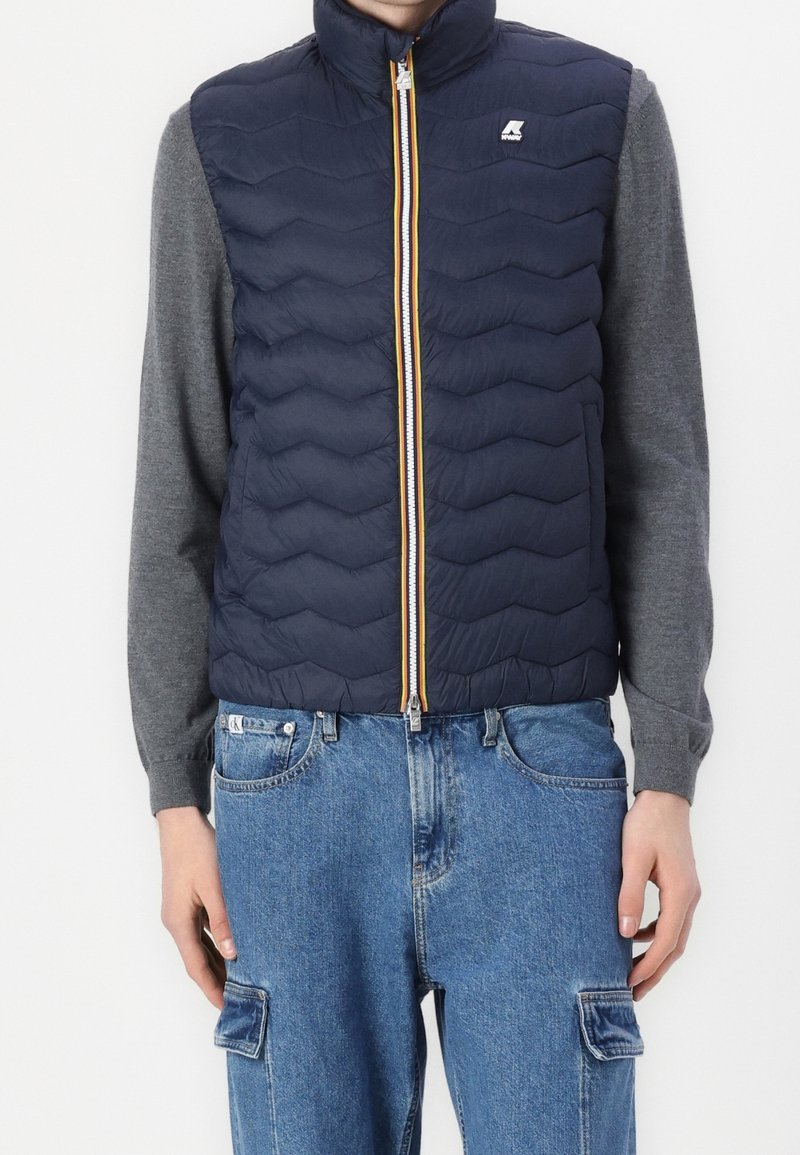 Person som bär en marinblå kviltad väst med framdragkedja och logotyp, grå långärmad tröja och blå denim cargo-jeans, stående mot vit bakgrund.
