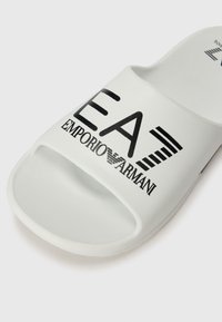 Bijela klizna sandala od gume s otvorenom petom. Sadrži crni otisnut logo "EA7 EMPORIO ARMANI" i dijagonalne pruge. Glatka tekstura.