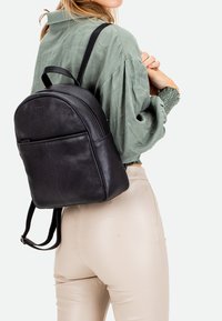 Zaino in pelle nera con parte superiore arrotondata, chiusura con zip, tasca frontale e spallacci regolabili, indossato sopra una maglia verde.