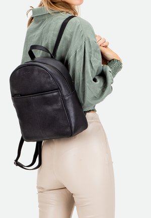 Zwarte leren rugzak met een afgeronde bovenkant, ritssluiting, voorzak en verstelbare banden, gedragen over een groene top.