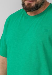 Zielona koszulka z krótkim rękawem, wykonana z miękkiego materiału. Posiada okrągły dekolt oraz małe logo w górnym lewym rogu.