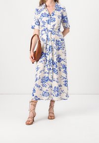 Žena v bílé midi šaty s modrým květinovým vzorem, v béžových sandálech na podpatku se šněrováním a držící hnědou kabelku přes rameno.