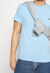 Světle modré bavlněné tričko s krátkým rukávem, doplněné šedou mikinou přehozenou přes rameno. Volný střih a jednoduchý design.