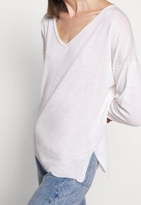 Hellrosa, V-Ausschnitt, langärmeliger Pullover mit lockerer Passform, strukturiertem Stoff und seitlichen Schlitzen; kombiniert mit blauen Jeans.