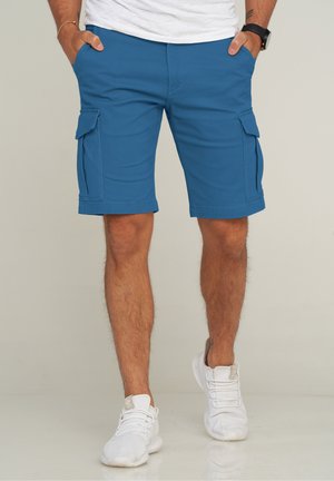 Blaue Cargo-Shorts mit glatter Textur, ausgestattet mit Seitentaschen und geradem Schnitt. Getragen mit weißen Sportschuhen.