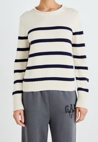 Kremowy sweter w granatowe paski, dzianinowa faktura, okrągły dekolt, ściśnięte wykończenia na dole i rękawach. W połączeniu z szarymi spodniami dresowymi z nadrukowanym logo.
