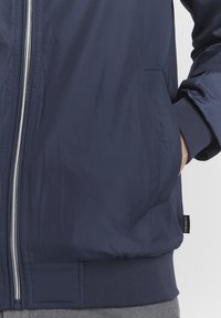 Jachetă de bombardier marină, dintr-un material neted, cu fermoar frontal, manșete ribbed și buzunare laterale. Eticheta este vizibilă pe partea stângă.