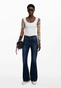 Tæt siddende hvid top med mønstrede lange ærmer, kombineret med vidde  mørk denim jeans. Modellen holder en lille brun taske med farverige detaljer.