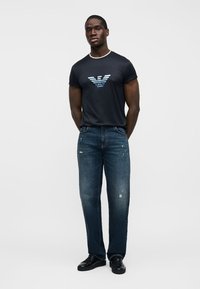 Άνδρας που στέκεται με τα χέρια πίσω από την πλάτη, φοράει μπλε σκούρο μπλουζάκι με λογότυπο, ξεθωριασμένο μπλε τζιν και μαύρα αθλητικά παπούτσια πάνω σε λευκό φόντο.