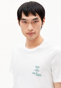 Weißes Baumwoll-T-Shirt mit einem grünen Druckdesign, "Gemacht, um einen Unterschied zu machen", auf der linken Brustseite.