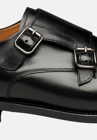 Schwarzer Lederschuh mit glatter Oberfläche, ausgestattet mit einem Riemen und doppelten Schnallen. Gekurvtes Design und verstärkte Nähte sind sichtbar.