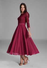 Robe à corsage en dentelle bourgogne avec des manches trois-quarts ; jupe ample et fluide en chiffon ; accompagnée de sandales à talons hauts avec des accents dorés.