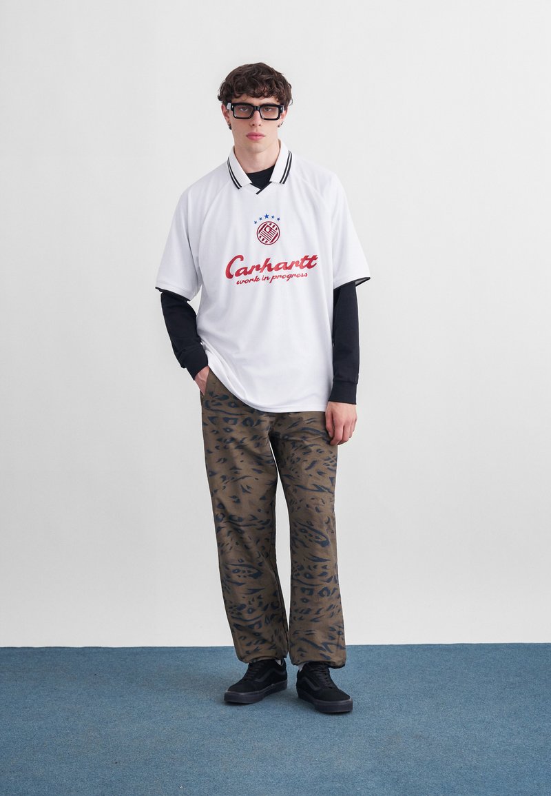 Junger Mann, der ein übergroßes weißes Carhartt-Polo-Shirt über schwarzem Langarmshirt, braune gemusterte Hose und schwarze Sneakers trägt und auf einem blauen Teppich steht.