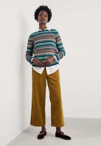 Knitted green and multicolored striped sweater layered over a white collared shirt, paired with wide-legged mustard corduroy pants and dark shoes.