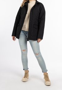 Svart quiltad jacka med avslappnad passform, två framfickor, tryckknappsstängning, bärs över en beige tröja och slitna ljusblå jeans.
