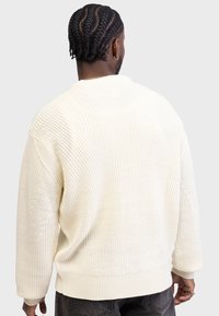 Jersey de punto grueso en color crema con textura acanalada, corte holgado, mangas anchas y hombros caídos, que presenta un diseño de costuras en la parte trasera.