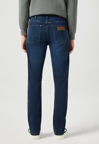 Donkerblauwe spijkerbroek met rechte pijpen, versterkte naden, twee achterzakken en een leren bruine patch op de tailleband.