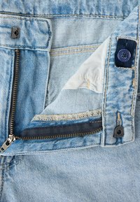 Ανοιχτό μπλε τζιν από denim με ανοιχτό μπρούτζινο φερμουάρ, ορατό κουμπί, ραφές και μέρος του εσωτερικού λευκού υφάσματος της τσέπης.