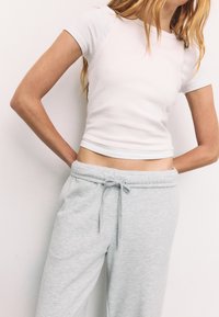 Donna che indossa una maglietta aderente bianca a maniche corte e pantaloni della tuta grigio chiaro con cordino, in piedi contro un muro bianco semplice con le mani dietro la schiena.