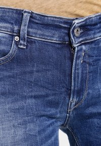 Jeans de mezclilla azul oscuro con un acabado desgastado. Presentan un cierre de botón y cremallera, diseño de cinco bolsillos y detalles de costura visibles.