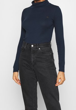 Femme portant un col roulé bleu marine à manches longues rentré dans un jean noir taille haute avec boutons et poches visibles à l’avant.