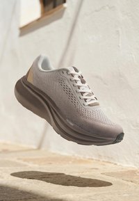 Chaussure de course gris clair avec lacets blancs, semelle épaisse marron foncé et empeigne en maille respirante, projetant une ombre sur le pavé en pierre.