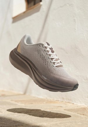 Hellgrauer Laufschuh mit weißen Schnürsenkeln, dicker dunkelbrauner Sohle und atmungsaktivem Strickobermaterial, wirft einen Schatten auf den Steinpflasterboden.