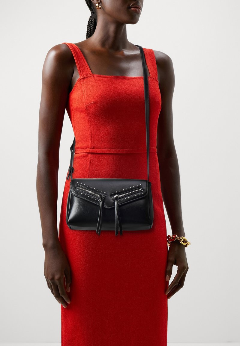 Kobieta w czerwonej sukience bez rękawów, z czarną, skórzaną torbą na ramię z dwoma kieszeniami na zamek i ozdobnymi ćwiekami.