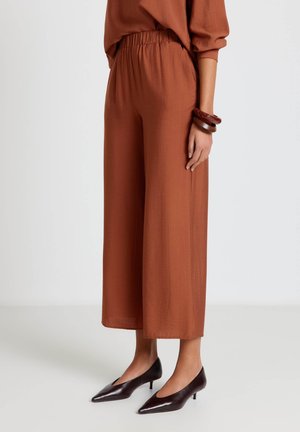 Donna che indossa pantaloni larghi color ruggine a gamba corta, top abbinato, scarpe scure marrone con tacco basso a punta e due braccialetti marroni spessi al polso.