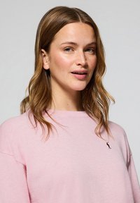 Ljusrosa tröja med rund halsringning, mjukt material, med en liten broderad logotyp på bröstet. Håret är vågigt och axellångt.