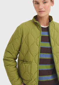 Gequilted groene jas met een staande kraag en een voorzak. De stof heeft een gladde textuur met gebogen stikselpatronen.