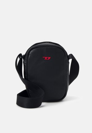 Sac bandoulière noir avec une large sangle et un petit logo rouge sur le devant, sur un fond blanc uni.