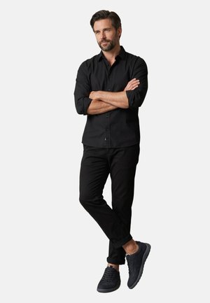 Muž s vousy, oblečený v černé košili na knoflíky, černé kalhoty a černé tenisky, stojící se zkříženýma rukama a jednou nohou přes druhou.