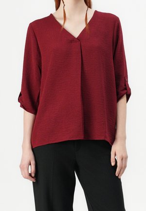 Femme portant un chemisier ample, texturé, rouge foncé avec des manches trois-quarts et un pantalon noir sur un fond blanc uni.