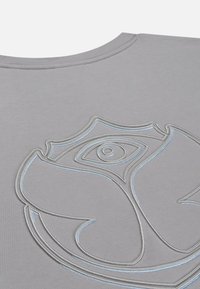 Close-up van een grijze stof met een geborduurd patroon van een gestileerde roos met een oog in het midden, omrand met lichtblauwe en grijze draad.