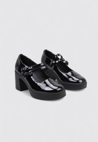 Zapatos Mary Jane de charol negro con tacón ancho, que presentan dos correas ajustables y una punta redonda. Acabado suave y brillante.