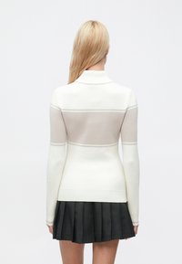 Hvit genser med en lys beige stripe over ryggen, med krage og tettsittende ermer. Kombinert med et plisssert mørkegrått skjørt.