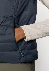 Colete acolchoado azul-marinho com textura acolchoada, apresentando um bolso lateral e uma bainha ajustável. Camisa de manga longa de cor clara por baixo.