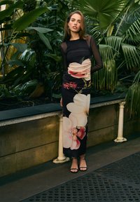 Czarna sukienka w kwiatowe wzory, z marszczeniami i długimi przezroczystymi rękawami. Noszona z czarnymi sandałami na cienkich paskach w bujnym, zielonym otoczeniu.