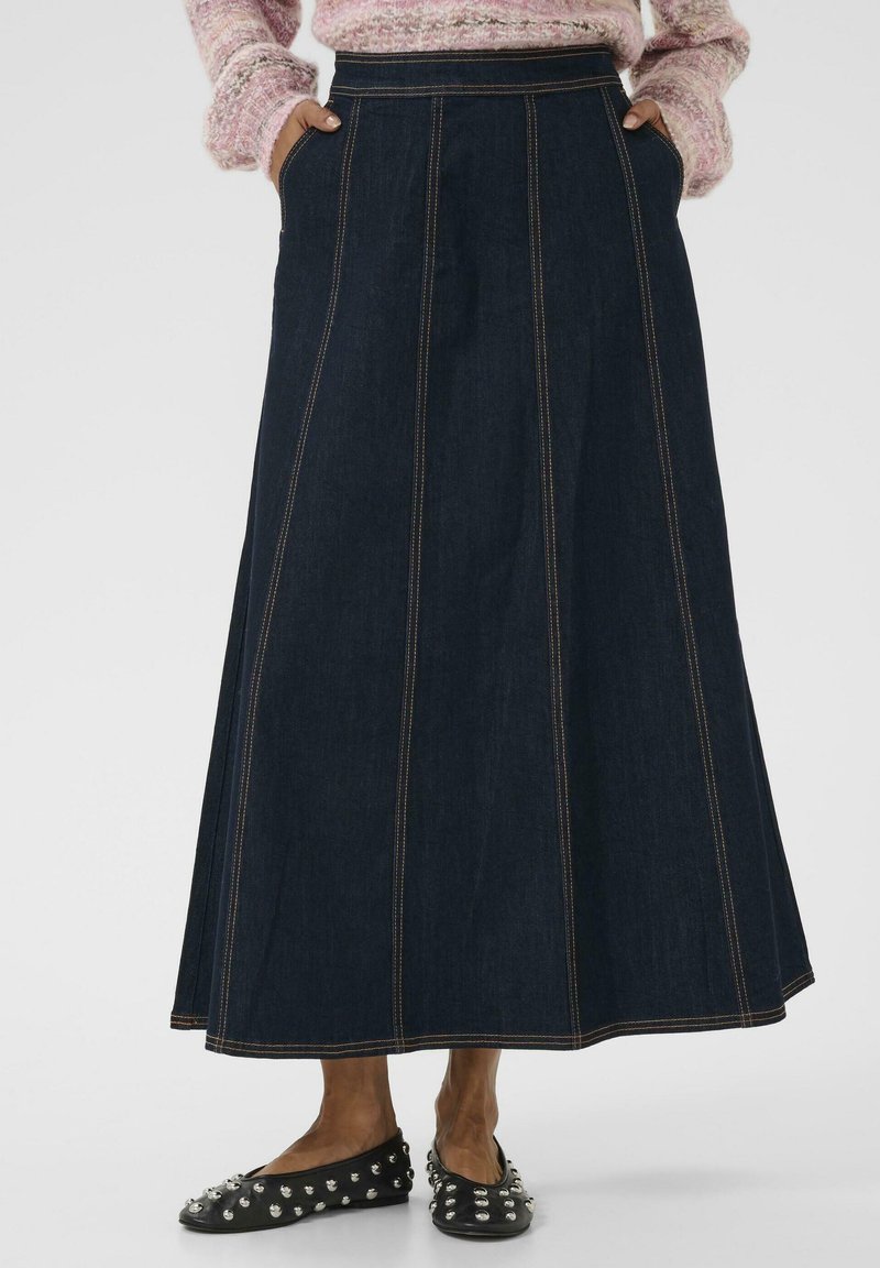 Jupe en denim longue et évasée de couleur bleu foncé avec des coutures visibles. Dotée de poches latérales et d'une texture lisse. Associée à des ballerines noires à clous.