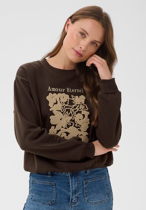 Donna con lunghi capelli castani inclina la testa, indossa una felpa marrone con "Amour Eternel" e motivo floreale beige, abbinata a jeans blu.