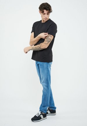 Giovane uomo con maglietta nera e jeans che guarda e tiene il suo avambraccio sinistro tatuato, in piedi contro uno sfondo chiaro e uniforme.