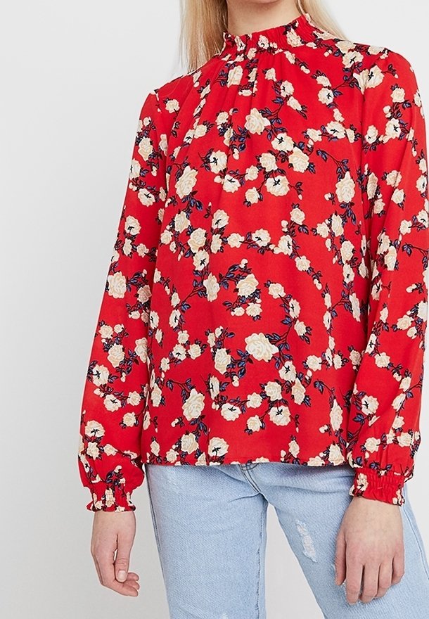 Femme portant un chemisier rouge à manches longues avec un imprimé floral blanc et des poignets à volants, associé à un jean déchiré bleu clair.