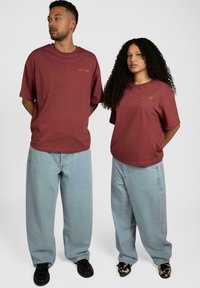 Burgunderfargede bomulls-T-skjorter med et lite gult grafikk; kombinert med lyseblå, løstsittende jeans. Sorte og mønstrete sko. Enkelt design.