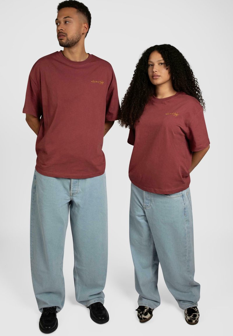 Burgunderfargede bomulls-T-skjorter med et lite gult grafikk; kombinert med lyseblå, løstsittende jeans. Sorte og mønstrete sko. Enkelt design.