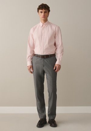 Giovane uomo in piedi che indossa una camicia rosa a righe con bottoni, pantaloni eleganti grigi, cintura marrone e scarpe in pelle marrone scuro contro una parete beige.